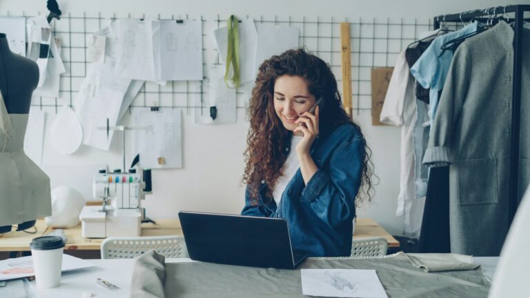 Créatrice indépendante souriante au téléphone devant son ordinateur portable, dans son atelier de couture